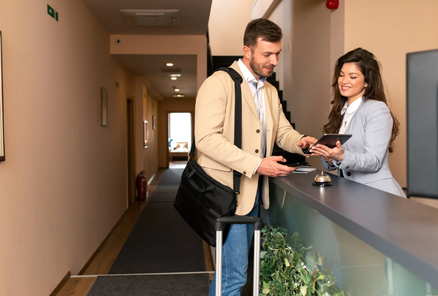 Young business man check-in in extended stay hotel market, smiling female receptionist behind the hotel counter showing him available rooms on tablet.