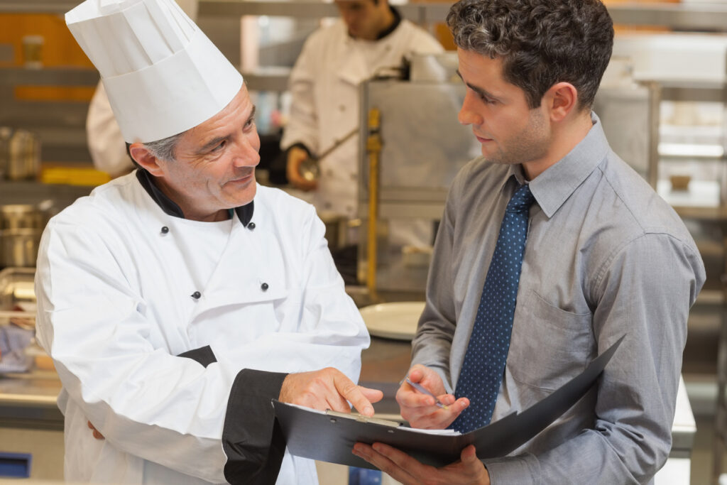 An older white man in a chef uniform shows a younger white man in a tie something on a clipboard. They're in an industrial kitchen.